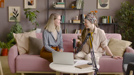 Two-Women-Recording-A-Podcast-Talking-Into-A-Microphone-Sitting-On-Sofa-In-Front-Of-Table-With-Laptop-And-Documents-1