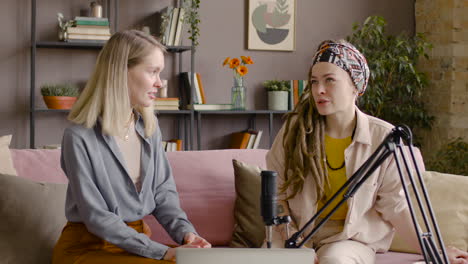 Two-Women-Recording-A-Podcast-Talking-Into-A-Microphone-Sitting-On-Sofa-In-Front-Of-Table-With-Laptop