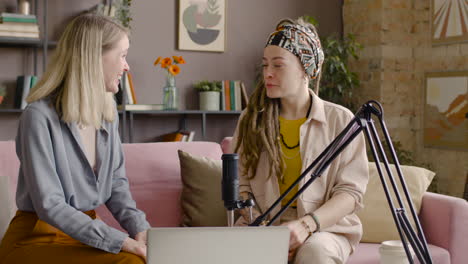 Two-Women-Recording-A-Podcast-Talking-Into-A-Microphone-Sitting-On-Sofa-In-Front-Of-Table-With-Laptop-1