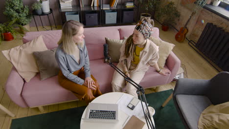 Top-View-Of-Two-Women-Recording-A-Podcast-Talking-Into-A-Microphone-While-Sitting-On-Sofa-In-Front-Of-Table-With-Laptop-And-Documents