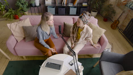 Top-View-Of-Two-Women-Recording-A-Podcast-Talking-Into-A-Microphone-While-Sitting-On-Sofa-In-Front-Of-Table-With-Laptop-And-Documents-1