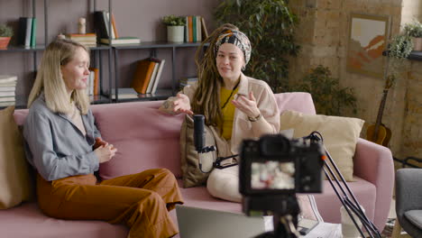 Rear-View-Of-A-Camera-Thats-Is-Recording-Two-Women-In-A-Podcast-Talking-Into-A-Microphone-While-They-Sitting-On-Sofa-1
