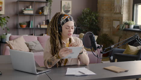 Woman-With-Dreadlocks-Recording-A-Podcast-Talking-Into-A-Microphone-Sitting-At-Desk-With-Laptop-And-Documents