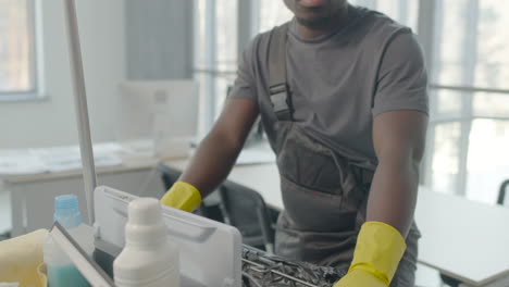 Cleaning-Man-Wearing-Gloves-And-Bandana-Posing-With-Cleaning-Cart-And-Looking-At-The-Camera-Inside-An-Office-1