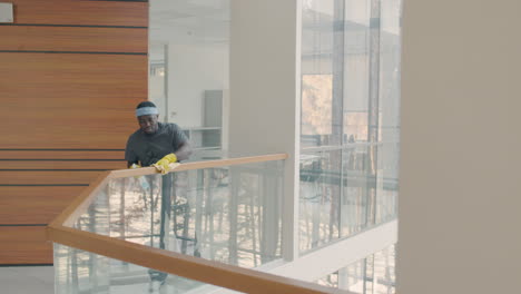 Distant-View-Of-Cleaning-Man-Wearing-Gloves-Cleaning-Stair-Railing-Inside-An-Office-Building