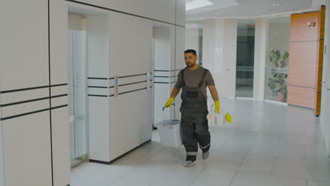 Arabischer-Reinigungsmann,-Der-Reinigungsmittel-Und-Wischeimer-Hält,-Während-Er-Einen-Korridor-In-Einem-Bürogebäude-Hinuntergeht