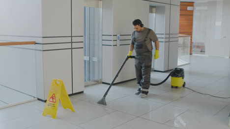 Arabic-Cleaning-Man-Wearing-Headphones-Cleaning-With-Pressure-Water-Machine-While-Dancing-Inside-An-Office-Building-Behind-A-Wet-Floor-Warning-Sign