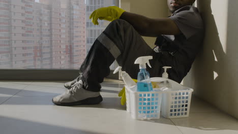 Close-Up-View-Of-Cleaning-Man-Sitting-On-The-Floor-And-Leaning-On-The-Wall-Near-A-Window-While-Looking-At-Camera-Inside-An-Office-Building