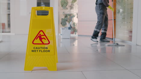 Cleaning-Man-Cleaning-The-Floor-With-Mop-Inside-An-Office-Building-Behind-A-Wet-Floor-Warning-Sign