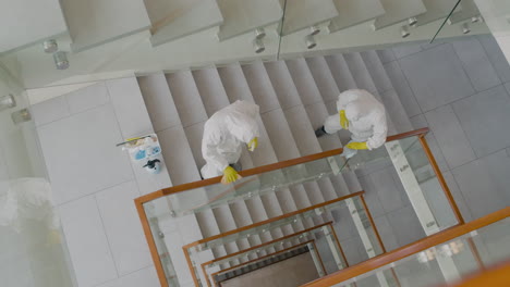Top-View-Of-Two-Cleaning-Men-Wearing-Personal-Protective-Equipment-Cleaning-Stair-Railings-Inside-An-Office-Building