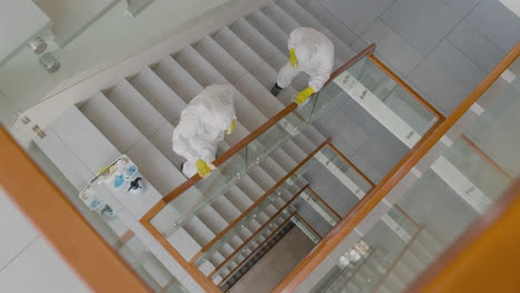 Top-View-Of-Two-Cleaning-Men-Wearing-Personal-Protective-Equipment-Cleaning-Stair-Railings-Inside-An-Office-Building-1