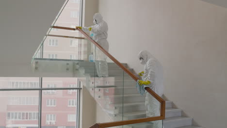 Rear-View-Of-Two-Cleaning-Men-Wearing-Personal-Protective-Equipment-Cleaning-Stair-Railings-Inside-An-Office-Building