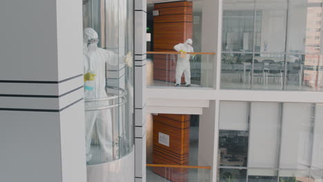 Distant-View-Of-Two-Cleaning-Men-Wearing-Personal-Protective-Equipment-Cleaning-Stair-Railings-And-Glass-Elevator-Inside-An-Office-Building