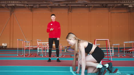 Two-Multiethnic-Female-Athletes-Taking-Position-On-The-Starting-Line-And-Then-Running-On-An-Indoor-Track-While-A-Male-Coach-Measuring-Time-With-A-Stopwatch