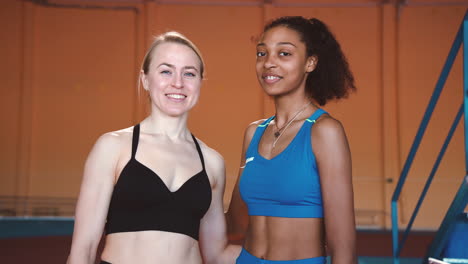 Porträt-Von-Zwei-Multiethnischen-Sportlerinnen,-Die-In-Die-Kamera-Lächeln,-Während-Sie-In-Einer-Indoor-Sportanlage-Stehen