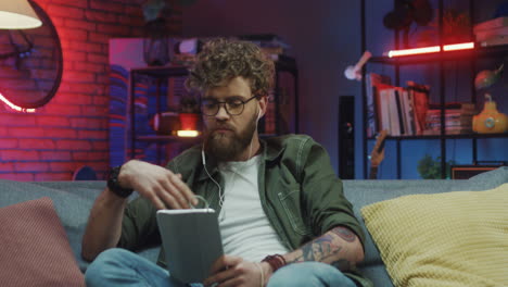 Young-Man-In-Glasses-And-Headphones-Sitting-On-Couch-With-A-Tablet-Watching-Something-Or-Listening-To-Music-At-Night