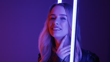 Close-Up-View-Of-Blonde-Young-Woman-Standing-Among-Neon-Lights-And-Smiling-Cheerfully-To-The-Camera