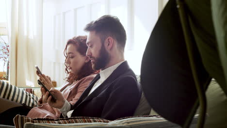Woman-And-Man-In-Formal-Wear-Using-Mobile-Phone-And-Talking-Together-While-Sitting-On-Sofa