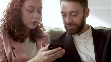 Close-Up-Of-Woman-And-Man-In-Formal-Wear-Using-Mobile-Phone-And-Talking-Together-While-Sitting-On-Sofa-1