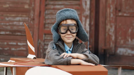 Portrait-Of-A-Cute-Little-Boy-With-Red-Hair-In-Hat-And-Glasses-Sitting-Outdoor-At-Wooden-Model-Of-Plane-And-Smiling-At-The-Camera