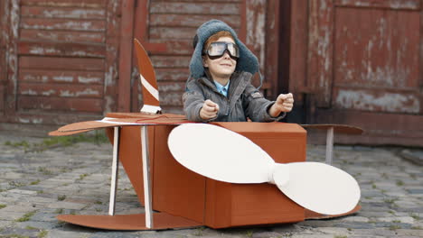 Glücklicher-Kleiner-Junge-Mit-Roten-Haaren-In-Hut-Und-Brille,-Der-Draußen-In-Einem-Hölzernen-Spielzeugmodell-Eines-Flugzeugs-Sitzt-Und-Davon-Träumt,-Flieger-Zu-Werden