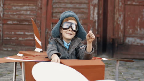 Happy-Little-Boy-With-Red-Hair-In-Hat-And-Glasses-Sitting-Outdoor-In-Wooden-Toy-Model-Of-Airplane-And-Dreaming-To-Be-Aviator-1