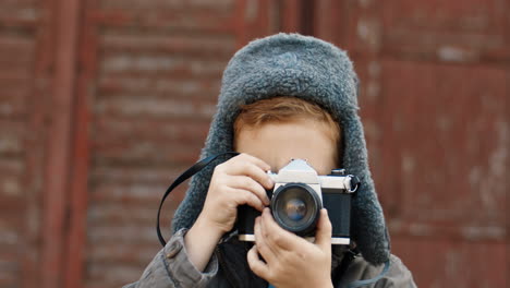 Portrait-Of-A-Cute-Little-Red-Haired-Boy-In-Aviator-Hat-Taking-Photos-With-Vintage-Camera-Outdoors-On-A-Wooden-Wall-Background