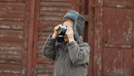 Cute-Little-Red-Haired-Boy-In-Aviator-Hat-Taking-Photos-With-Vintage-Camera-Outdoors-On-A-Wooden-Wall-Background