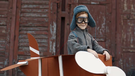Portrait-Of-A-Cute-Little-Red-Haired-Boy-In-Hat-And-Glasses-Standing-In-A-Cardboard-Plane-And-Smiling-At-The-Camera