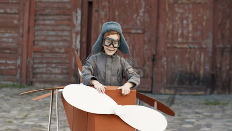 Cute-Little-Red-Haired-Boy-In-Hat-And-Glasses-Walking-Outdoors-In-A-Cardboard-Plane-Dreaming-To-Be-An-Aviator-1