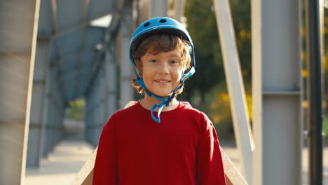 Portrait-Of-A-Cute-Little-Boy-In-Helmet-And-Red-Sweater-Looking-At-The-Camera-While-Standing-On-A-Bridge-On-A-Sunny-Day