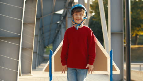 Portrait-Of-A-Cute-Little-Boy-In-Helmet-And-Red-Sweater-Looking-At-The-Camera-While-Standing-On-A-Bridge-On-A-Sunny-Day-1