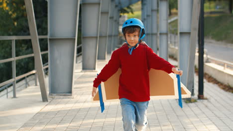Cute-Little-Boy-In-Helmet-And-Red-Sweater-With-Cardboard-Airplane-Wings-Running-On-A-Bridge-On-A-Sunny-Day