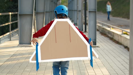 Back-View-Of-A-Little-Boy-In-Helmet-And-Red-Sweater-With-Cardboard-Airplane-Wings-Walking-Across-The-Bridge-And-Playing-As-A-Pilot