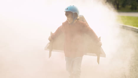 Happy-Little-Boy-Wearing-Cardboard-Airplane-Wings-With-Artificial-Smoke-Behind-Running-In-The-Park-And-Playing-As-A-Pilot