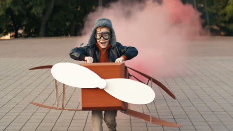 Cute-Little-Red-Haired-Boy-In-Hat-And-Glasses-Running-In-The-Park-Holding-A-Cardboard-Plane-With-Smoke-Behind-And-Playing-As-An-Aviator