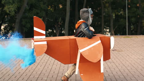 Side-View-Of-A-Cute-Little-Running-In-The-Park-Holding-A-Cardboard-Plane-With-Smoke-Behind-And-Playing-As-An-Aviator