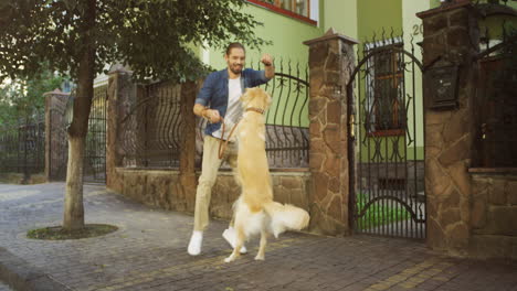 Junger-Mann,-Der-Mit-Seinem-Hund-In-Einer-Malerischen-Straße-Mit-Häusern-Spazieren-Geht,-Damit-Trainiert-Und-Spielt