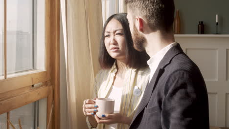 Man-In-Formal-Wear-And-Young-Woman-Holding-Mugs-And-Talking-Together-While-Standing-Near-The-Window-In-The-Morning