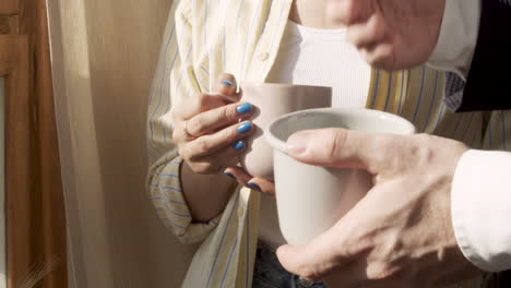 Close-Up-Of-Two-People's-Hands-Holding-Coffee-Cups-Near-The-Window-In-The-Morning