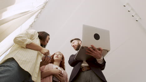Below-Shot-Of-A-Man-Holding-A-Laptop-Computer-And-Showing-Something-To-His-Female-Colleagues-While-Standing-In-The-Office-And-Talking-Together