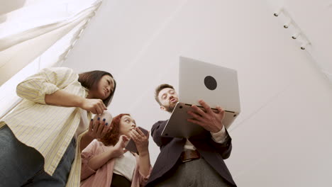 Below-Shot-Of-A-Man-Holding-A-Laptop-Computer-And-Showing-Something-To-His-Female-Colleagues-While-Standing-In-The-Office-And-Talking-Together-1