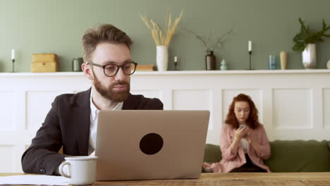 Bearded-Man-With-Glasses-Working-On-Laptop-Computer-While-A-Young-Woman-Using-Mobile-Phone-Sitting-On-Sofa