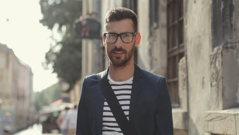 Portrait-Of-An-Attractive-Young-Man-In-Glasses-And-Stylish-Outfit-Turning-His-Head-To-The-Camera-And-Smiling-In-An-Old-Town-Street