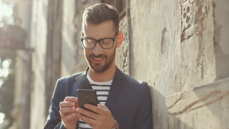Nahaufnahme-Eines-Lächelnden-Mannes-In-Stilvollem-Outfit,-Der-Sich-Auf-Der-Straße-An-Die-Wand-Lehnt-Und-Auf-Dem-Smartphone-SMS-Schreibt