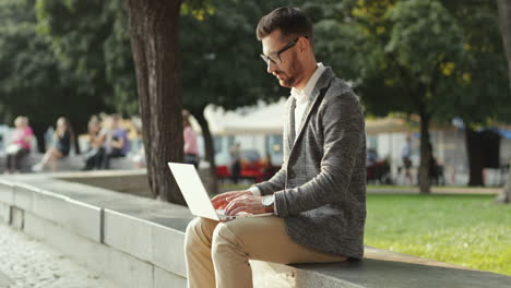 Handsome-Businessman-Sitting-On-Wall-In-The-City-Park-And-Working-On-His-Laptop-Computer