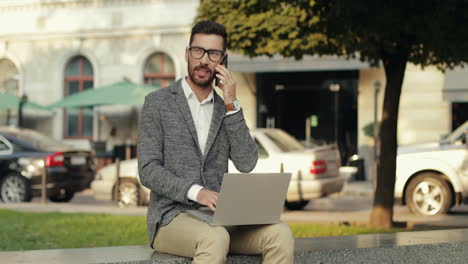 Good-Looking-Businessman-Talking-On-His-Mobile-Phone-While-Sitting-On-The-Wall-In-The-City-And-Working-On-His-Laptop-Computer-1