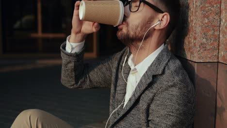Close-Up-Of-A-Relaxed-Businessman-Drinking-Coffee-And-Listening-Music-On-The-Smartphone-Via-Earphones-While-Sitting-On-Steps-Of-A-Building-In-The-City