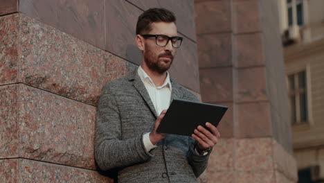 Stylish-Businessman-Standing-Outdoor-And-Leaning-Against-The-Wall-While-Tapping-And-Scrolling-On-The-Tablet-Device