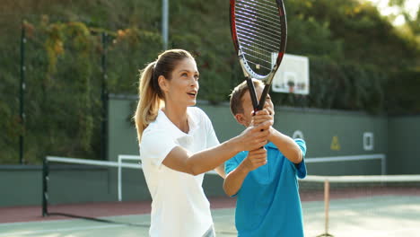 Smiling-Woman-Teaching-Her-Son-How-To-Hit-Ball-With-Racket-On-A-Tennis-Court